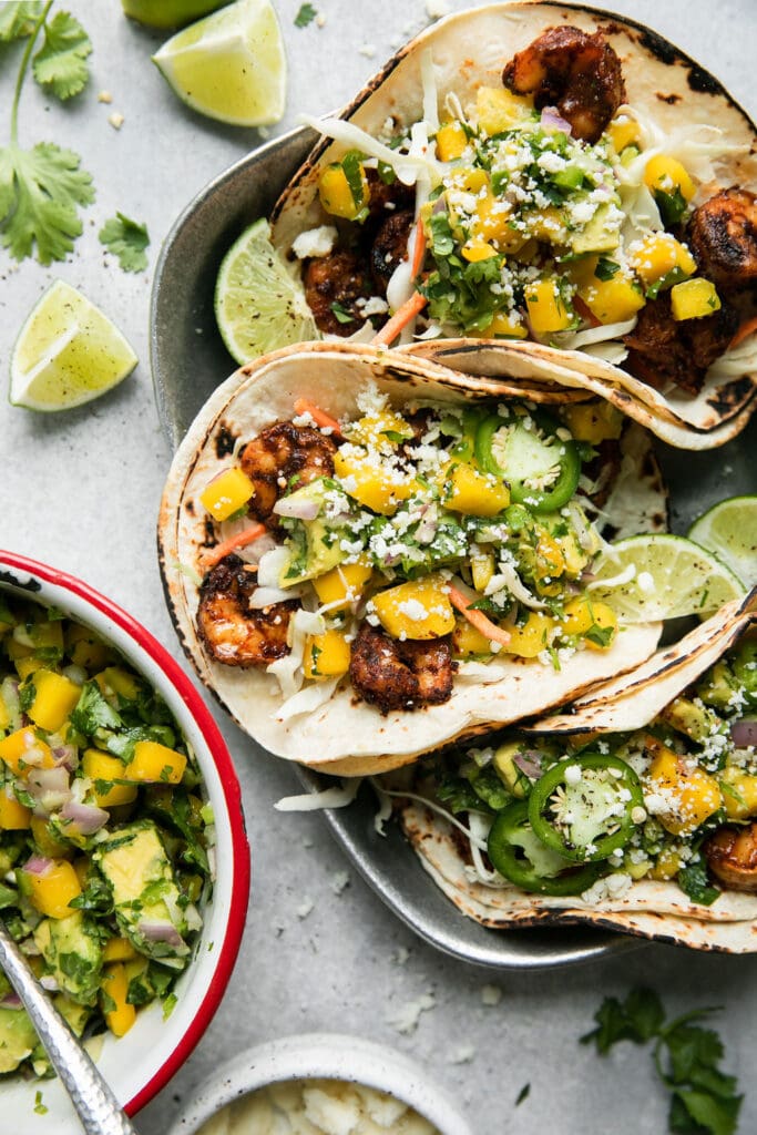 Three grilled shrimp tacos in charred corn shells topped with mango avocado salsa and cheese in a silver metal plate. 