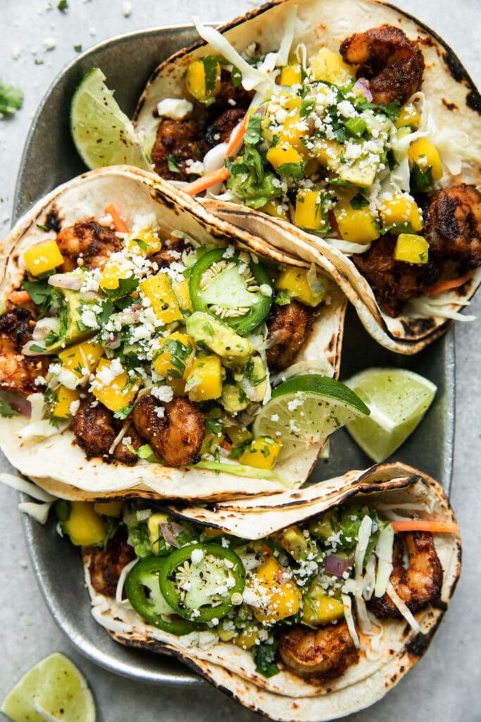 Overhead view of three grilled shrimp tacos in double layer charred tortillas topped with mango avocado salsa and cojita cheese in a metal serving tray.