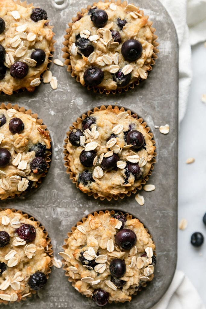 Overhead view blueberry oatmeal muffins with yogurt in muffin tin topped with blueberries and rolled oats.