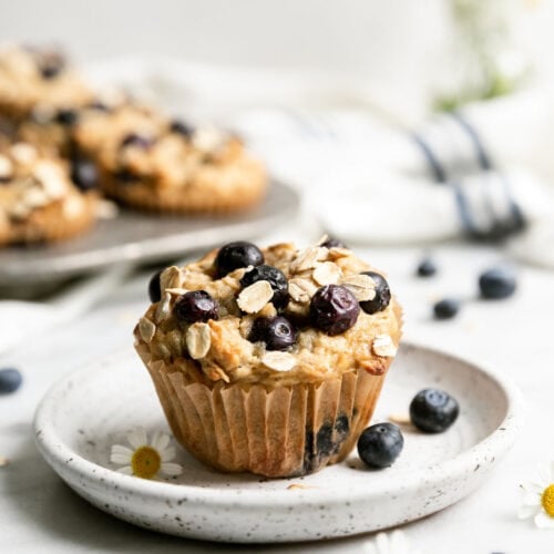 A single blueberry oatmeal muffin on white speckled plate, topped with juicy baked in blueberries.