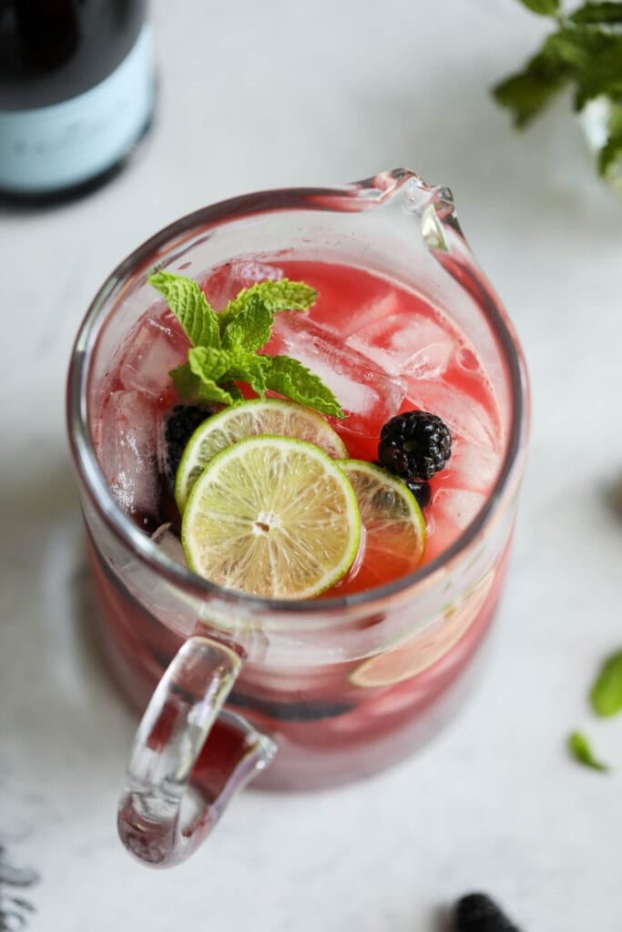 Overhead view looking into glass pitcher filled with blackberry lime prosecco cocktail with fresh lime slices floating on top.