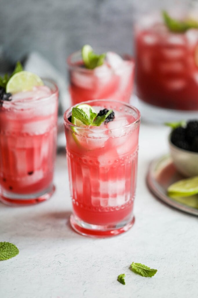 Tall glass filled with blackberry lime prosecco, garnished with fresh mint and lime slices, blackberries floating in drink. 