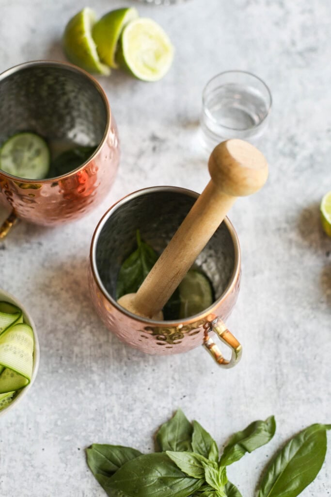 Overhead view copper cup with cucumber and basil being muddled with wooden muddler.