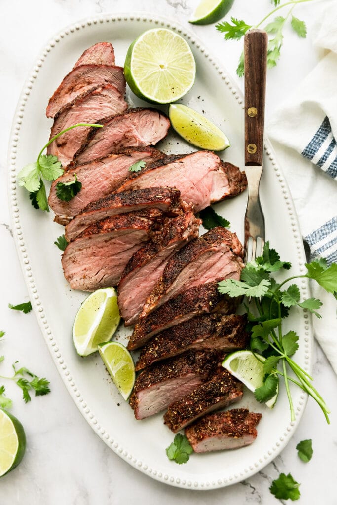Overhead view sliced grilled pork tenderloin on white platter with cilantro and lime wedges around. 