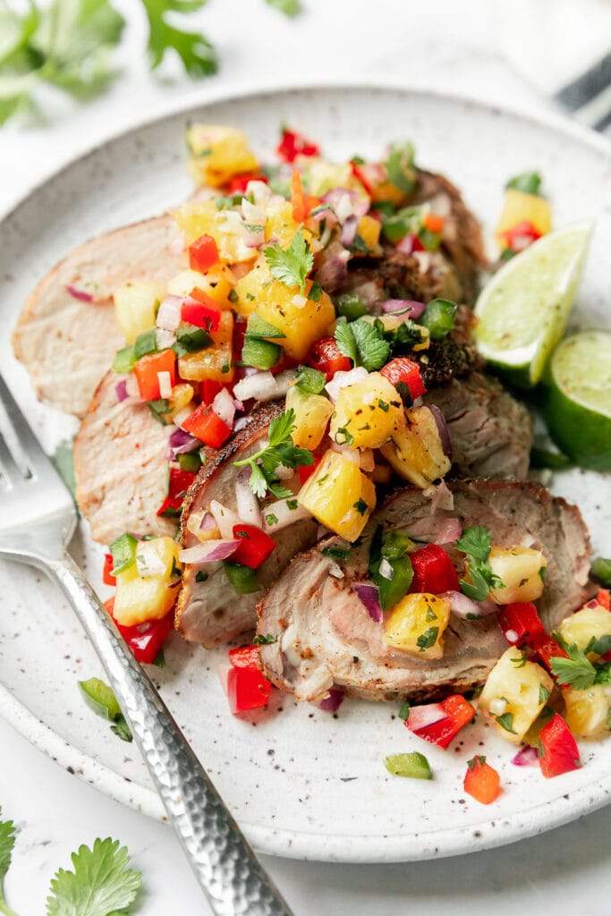 Grilled pork tenderloin cut into slices plated on white plate topped with homemade pineapple salsa, lime wedges on side.