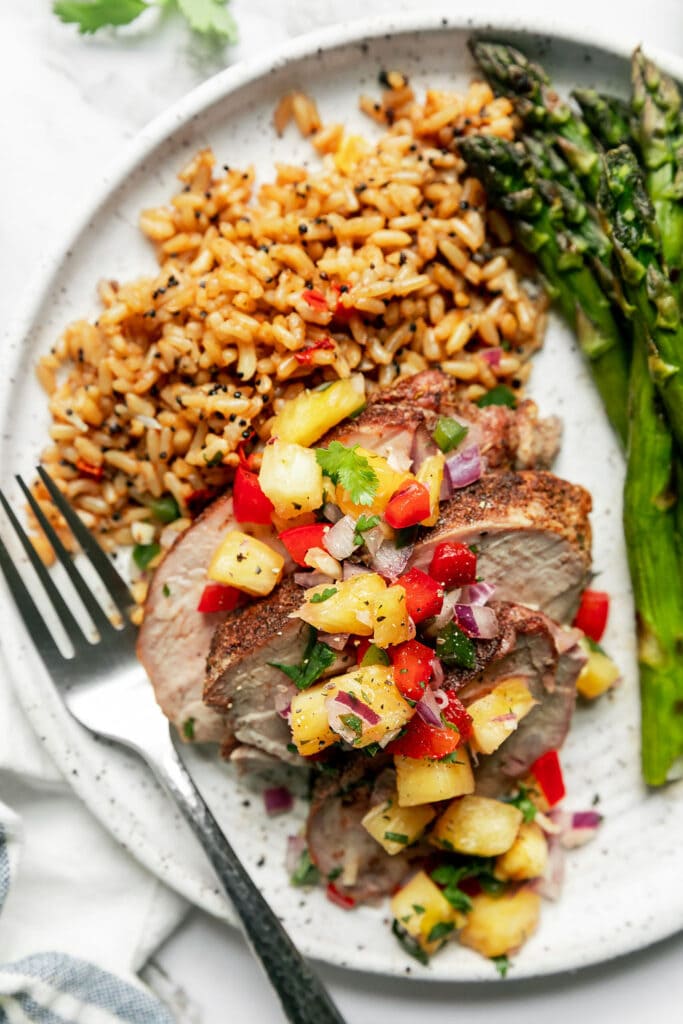 Grilled pork tenderloin sliced served on plate with rice pilaf and grilled asparagus, pineapple salsa topping grilled pork tenderloin slices. 
