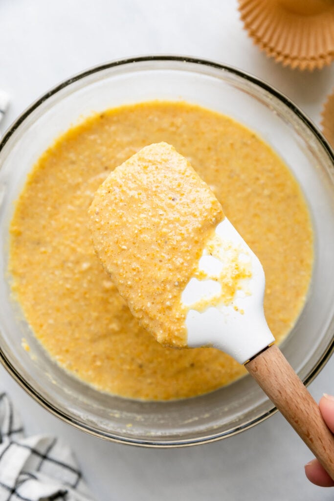 White spatula holding cornbread muffin batter over clear glass mixing bowl.