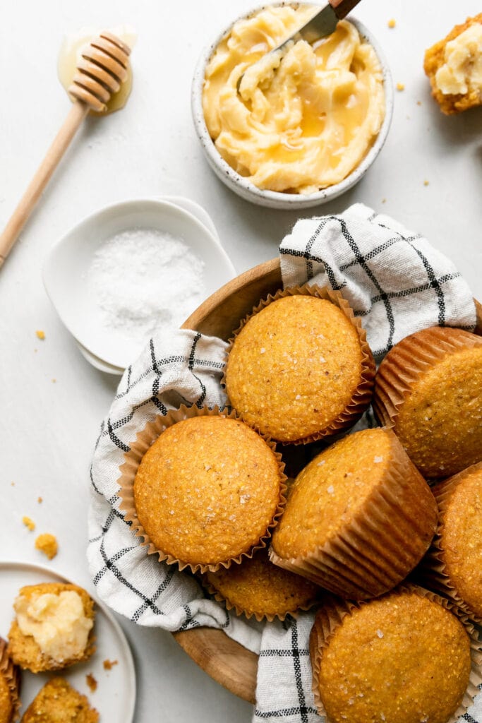 Liner covered wooden bowl filled with cornbread muffins sprinkled with sea salt.