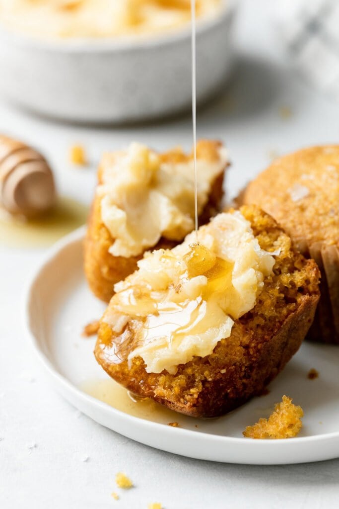 A cornbread muffin cut in half with thick spread of honey butter on cornbread muffin, honey drizzling over top. 