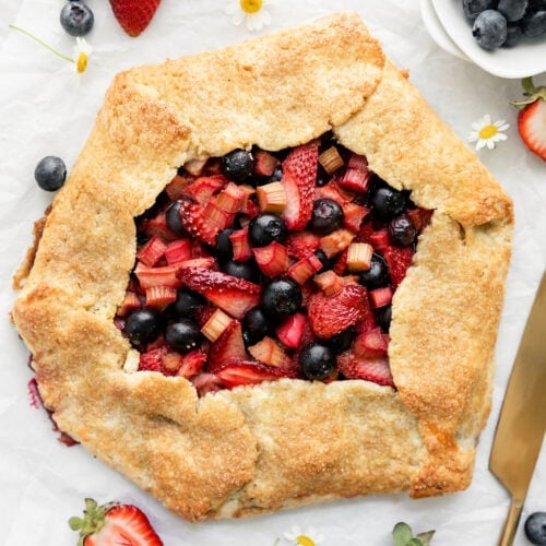 Overhead view strawberry blueberry galette with rhubarb in center of parchment paper, fresh berries and Shasta daisies scattered around.