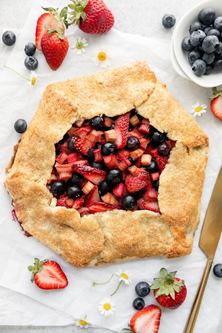 Overhead view of strawberry Galette with rhubarb surrounded by fresh fruit and fresh flowers. 