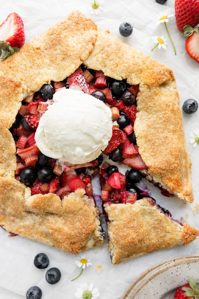 Overhead view strawberry blueberry galette with rhubarb, one slice cut and pulled back from entire pastry. Scoop of ice cream on center of galette.