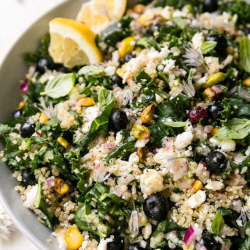 Quinoa kale salad with blueberries and feta in white bowl with lemon wedge on side.
