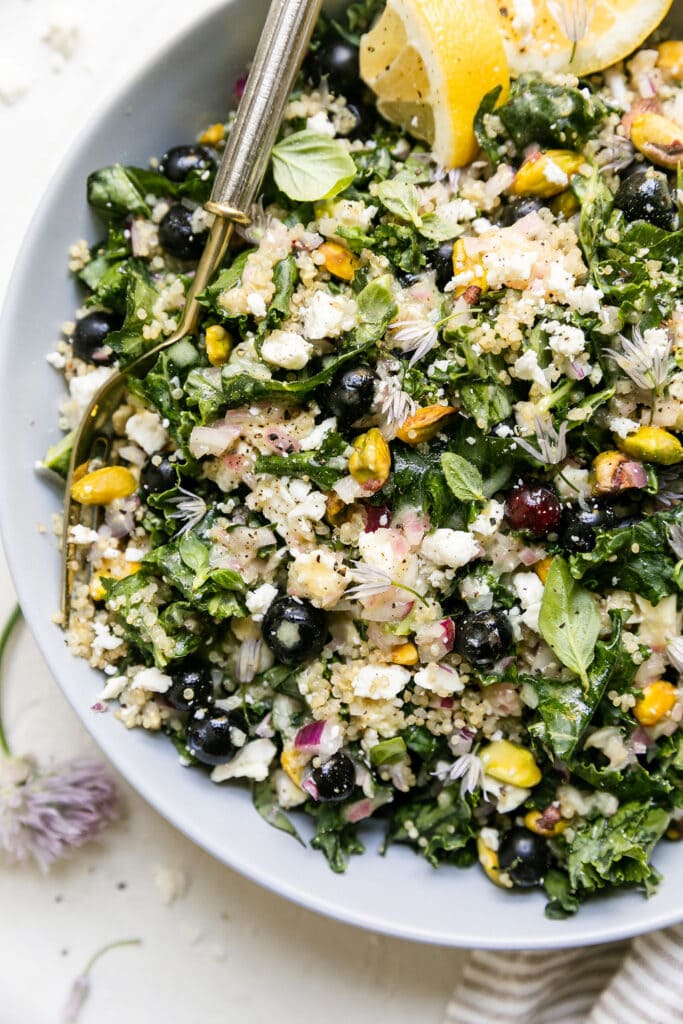 Close up overhead view quinoa kale salad with blueberries, feta, and pistachios in white bowl with gold fork.