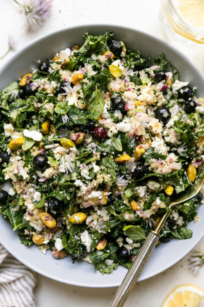 Overhead view quinoa kale salad with fresh blueberries, feta, and pistachios in white bowl. 