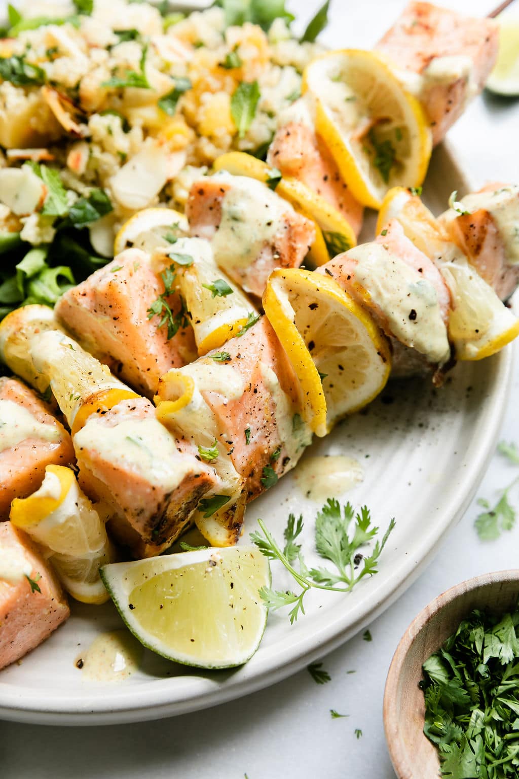 Close up view grilled salmon kabobs with lemon slices between salmon pieces, curry yogurt sauce drizzled on top. 