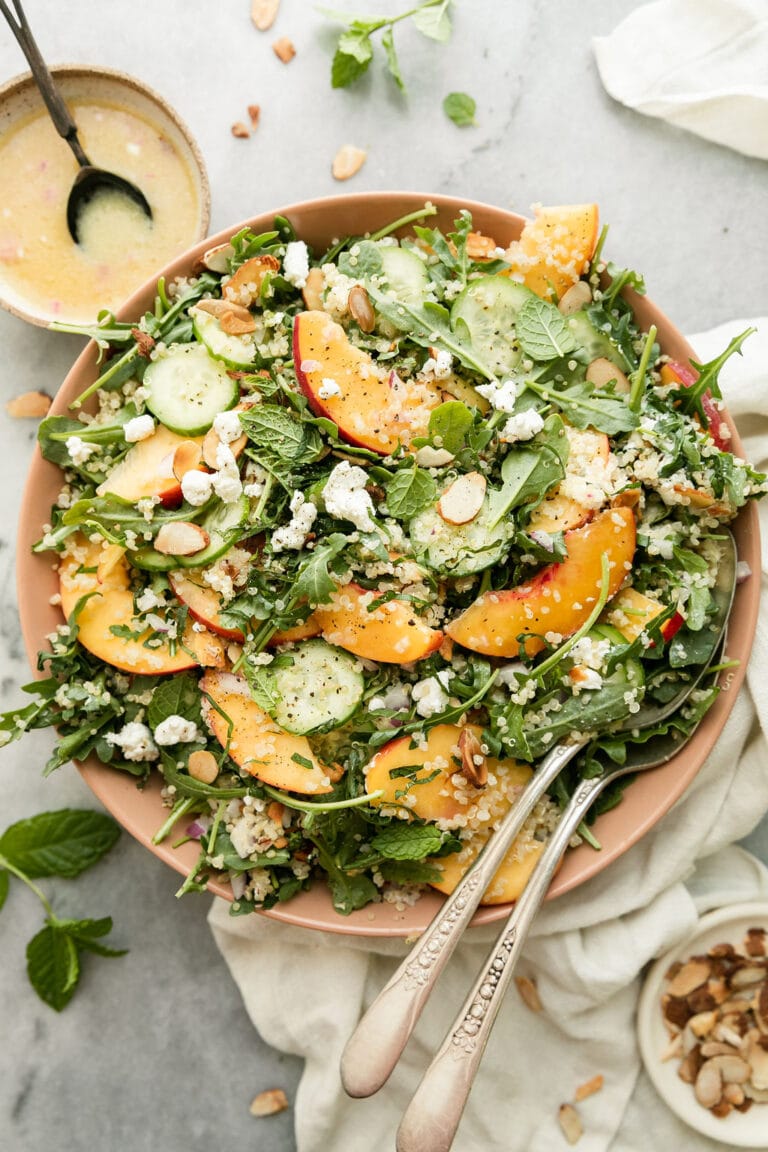A pink serving bowl filled with quinoa arugula salad with fresh peach slices, almond slices, and goat cheese.