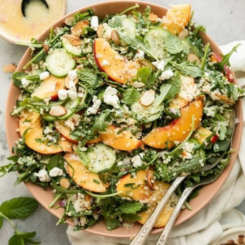 Overhead view quinoa arugula salad with peaches and goat cheese served in pink bowl.