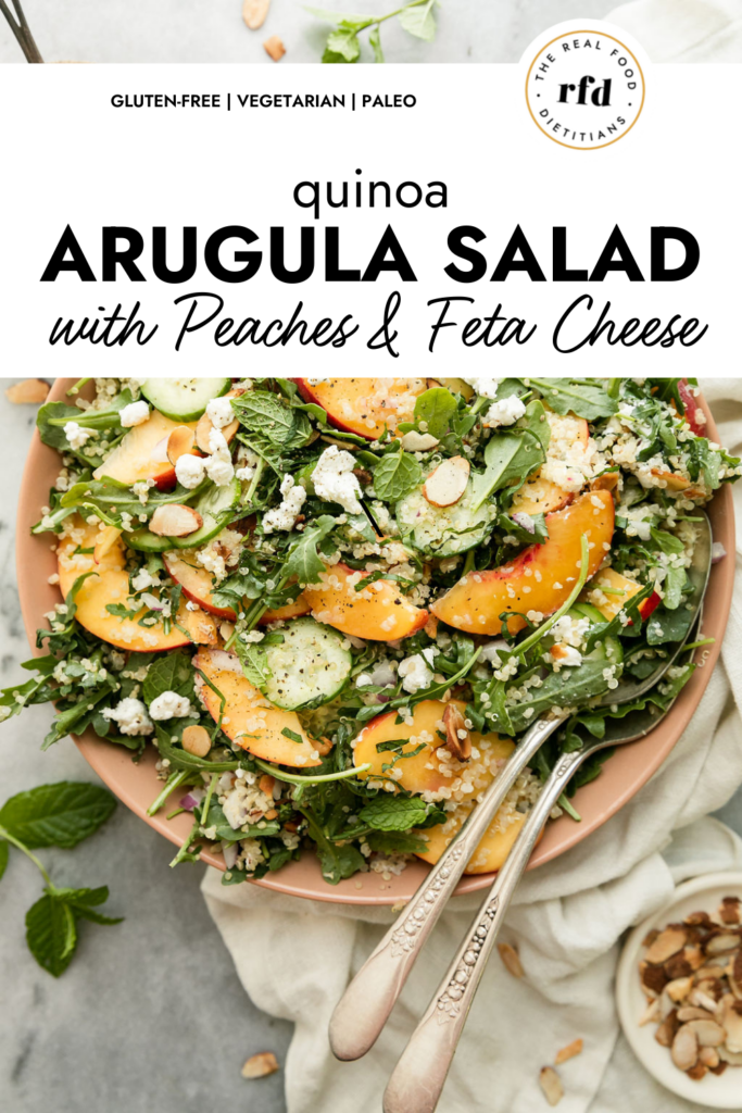 Quinoa arugula salad with peach slices and goat cheese crumbles in light pink bowl topped with toasted almonds.