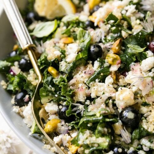 Closeup view quinoa kale salad with blueberries and feta crumbles in white bowl with gold fork.