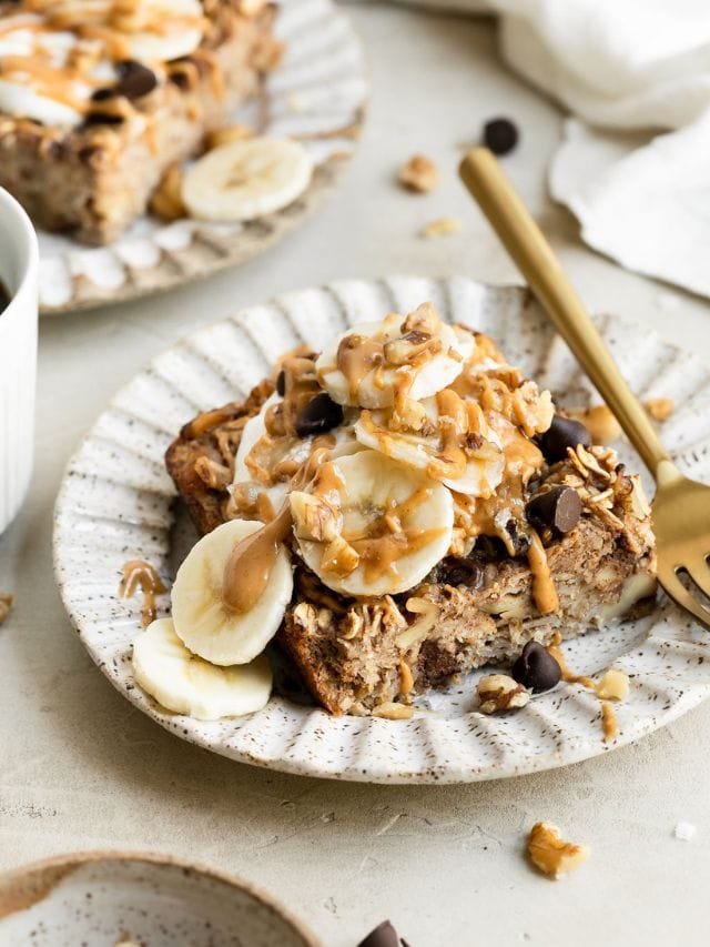 A serving of banana chocolate chip baked oatmeal on a plate topped with yogurt, banana slices, and peanut butter drizzle.