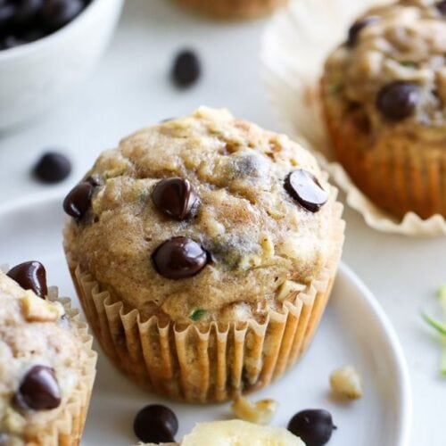 A single chocolate chip zucchini muffin topped with chocolate chips and walnuts on a white plate.