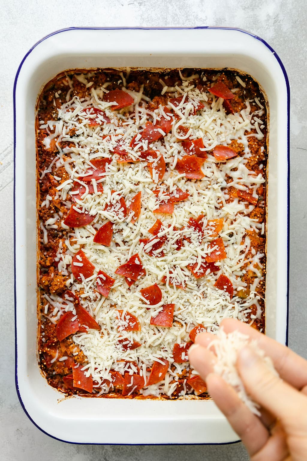 A hand sprinkling shredded mozzarella cheese over a pepperoni pizza quinoa casserole
