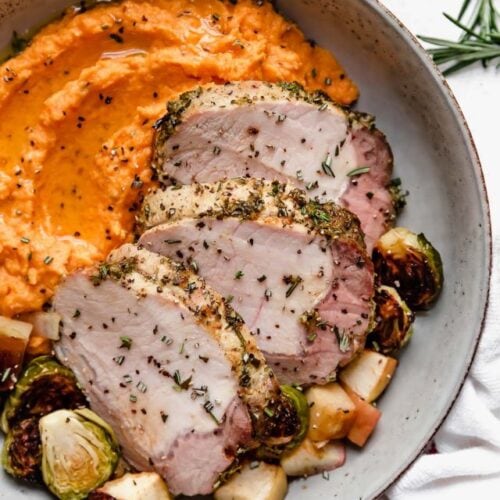 A shallow bowl filled with sliced pork loin, roasted Brussels sprouts and apples, and side of mashed sweet potatoes.