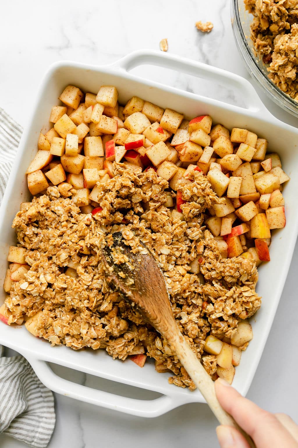 A wooden spoon spreading peanut butter topping over diced apples with cinnamon in white baking dish.