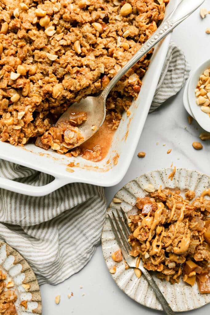 A silver spoon scooping up apple crumble with peanut butter topping from white baking dish.