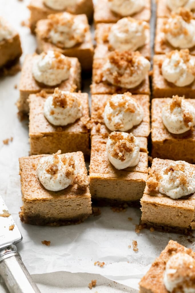 Several mini pumpkin cheesecake bars cut into squares topped with whipped cream and sprinkled with graham cracker crumble.
