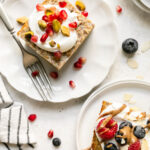 Protein baked oatmeal servings on white plates, topped with Greek yogurt, pomegranate seeds, pistachios, and fresh berries. 