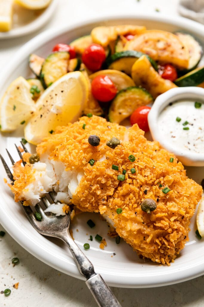 A crispy baked cod fillet on plate with fork full to show flaky texture, roasted vegetables on side.