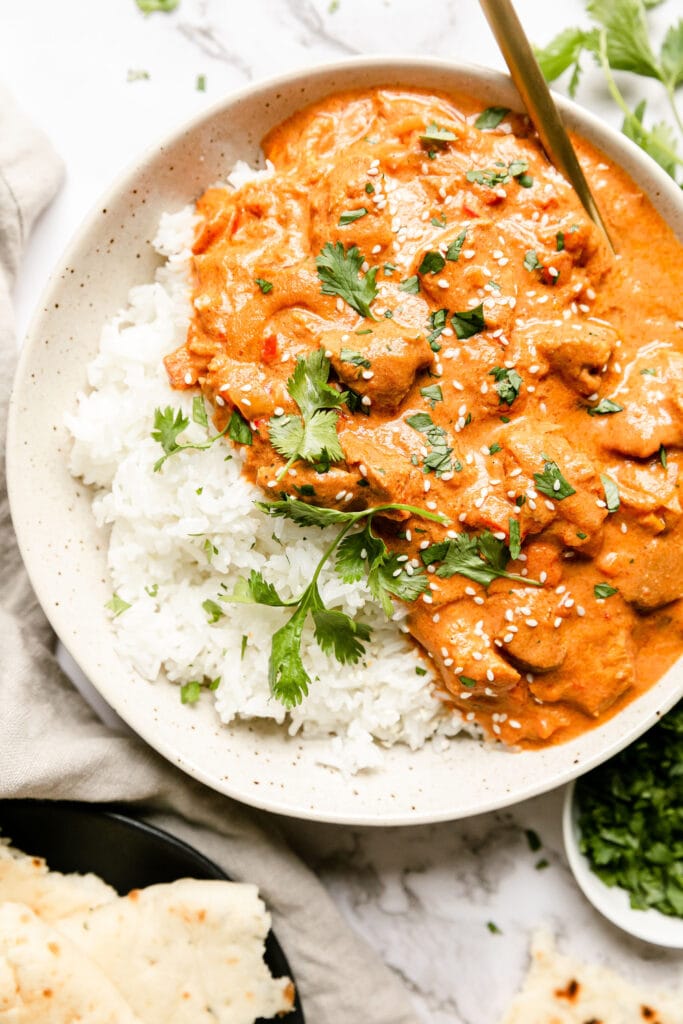 Bowl of creamy butter chicken on a plate with rice and cilantro on top. 