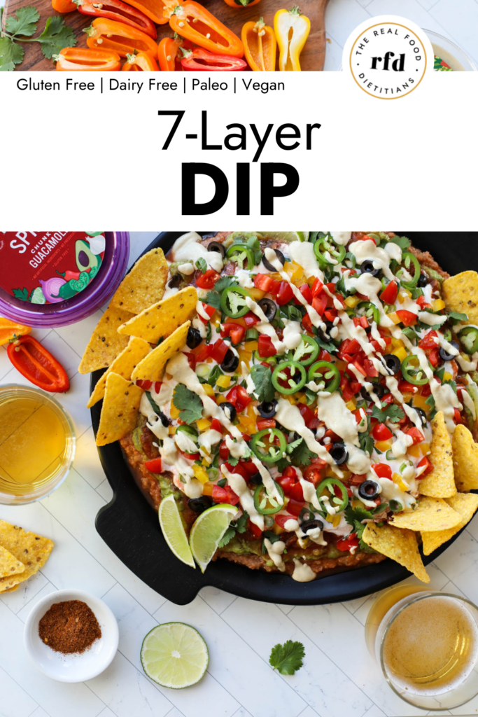 Overhead view 7 layer dip on a black round tray, topped with black olives, jalapeńo slices, and drizzled with plant based sour cream.