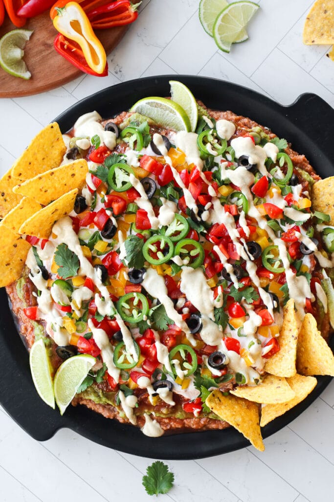 Overhead view 7 layer dip on black platter topped with fresh cut veggies, plant based sour cream, and jalapeńo slices. Corn tortillas sticking into top of dip.