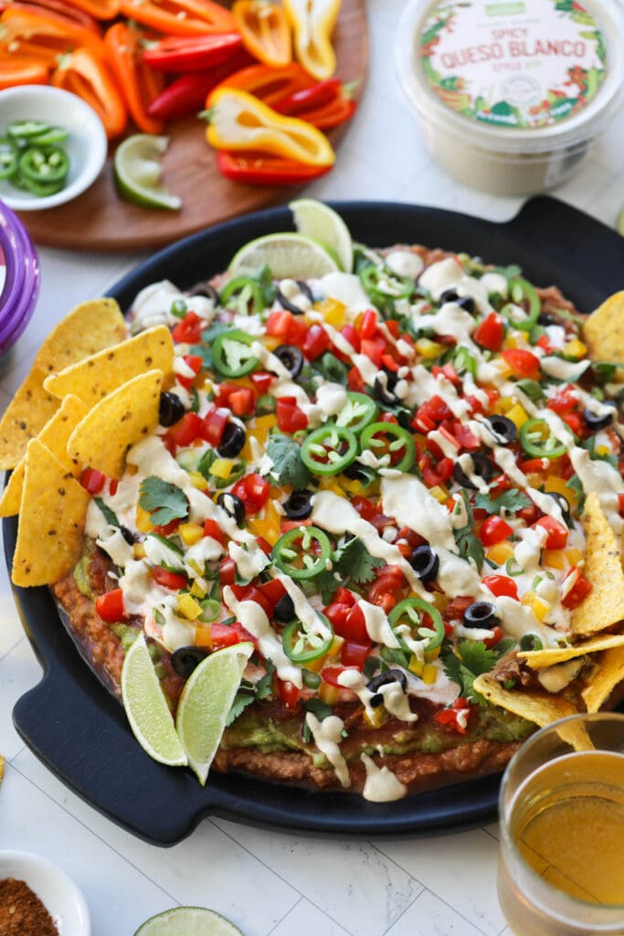 Overhead view 7 layer dip spread in layers on a black round tray. Topped with cut peppers, jalapeńo slices, black olives, and corn tortillas.