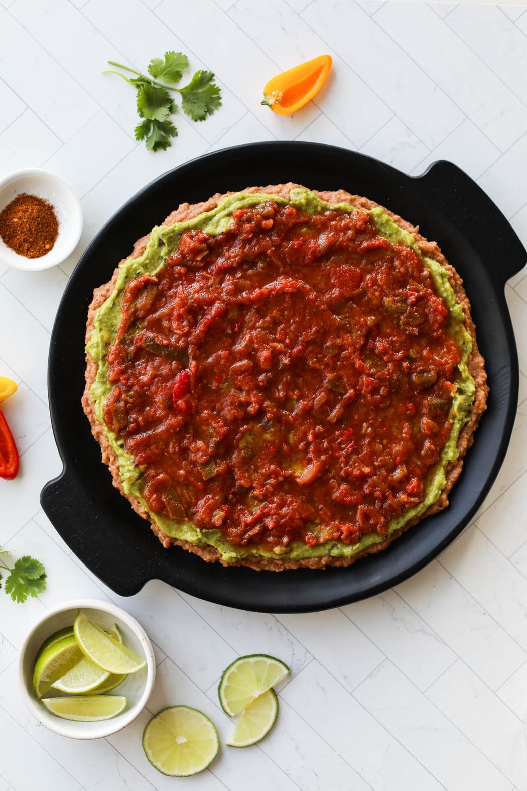Black platter with layers of refried beans, guacamole, and salsa spread in a circle.