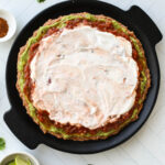 Round black tray with layers of refried beans, guacamole, salsa, and plant-based sour cream for 7 layer dip.