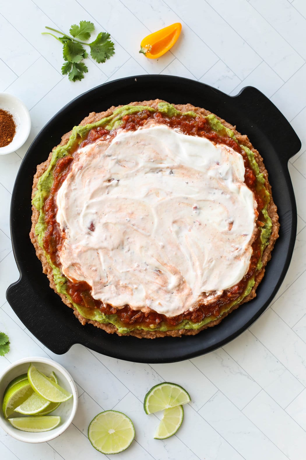 Black platter with layers of refried beans, guacamole, salsa, and sour cream spread in circle.