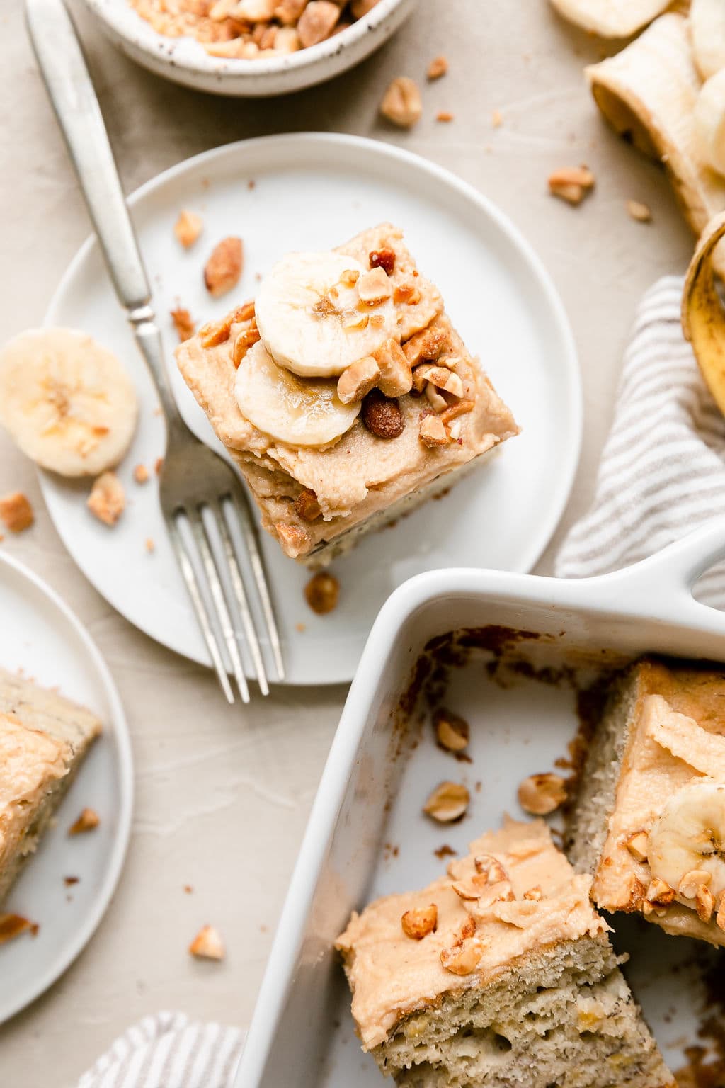 Overhead view piece of banana cake topped with frosting, banana slices, and chopped peanuts on a small white plate.