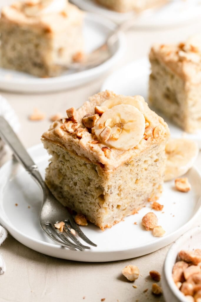 A square piece of fluffy banana cake topped with whipped peanut butter frosting with banana slices and chopped peanuts on a white plate.