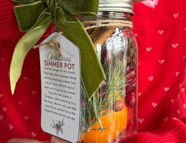 A woman in a red holiday sweater holding a glass mason jar filled with cranberries, orange, and cinnamon sticks. The jar has a pretty gift tag attached to it.