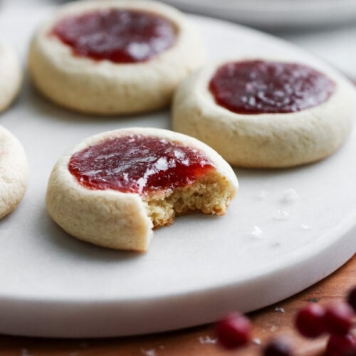 Raspberry Thumbprint Cookies on a marble round tray, front cookie has bite taken out.