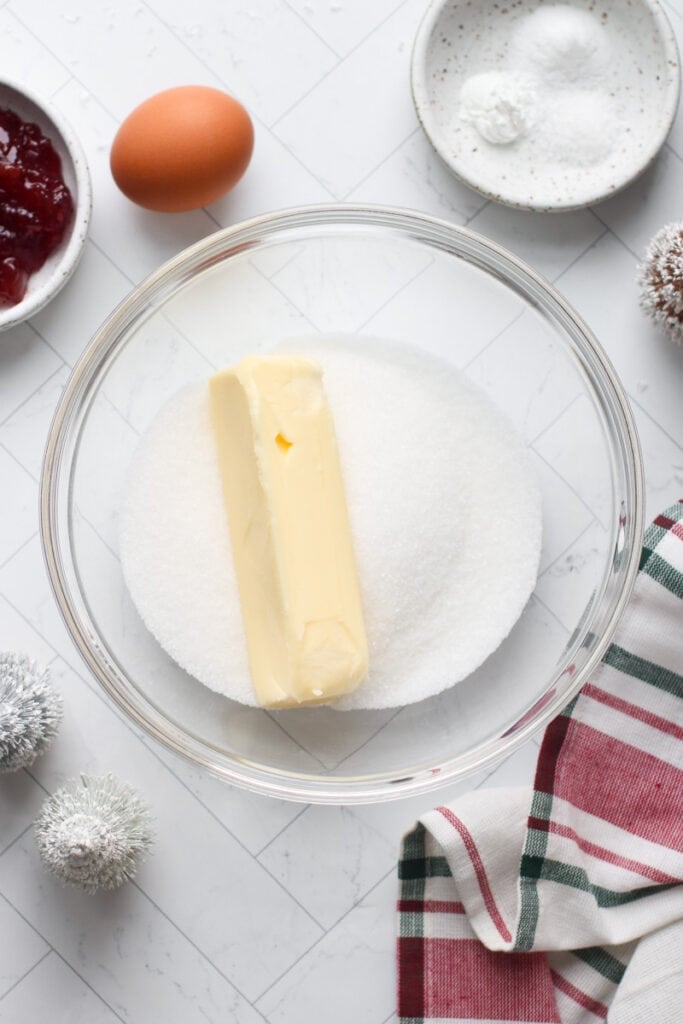 Sugar and a stick of butter in a clear glass mixing bowl for thumbprint cookies.