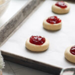 Thumbprint cookies lined up on parchment-covered baking sheet, filled with raspberry filling.