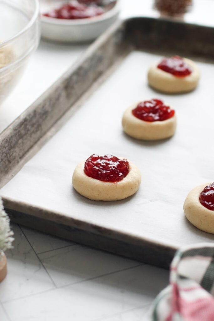 Side view thumbprint cookies filled with raspberry jam lined up on parchment-lined bakign sheet.