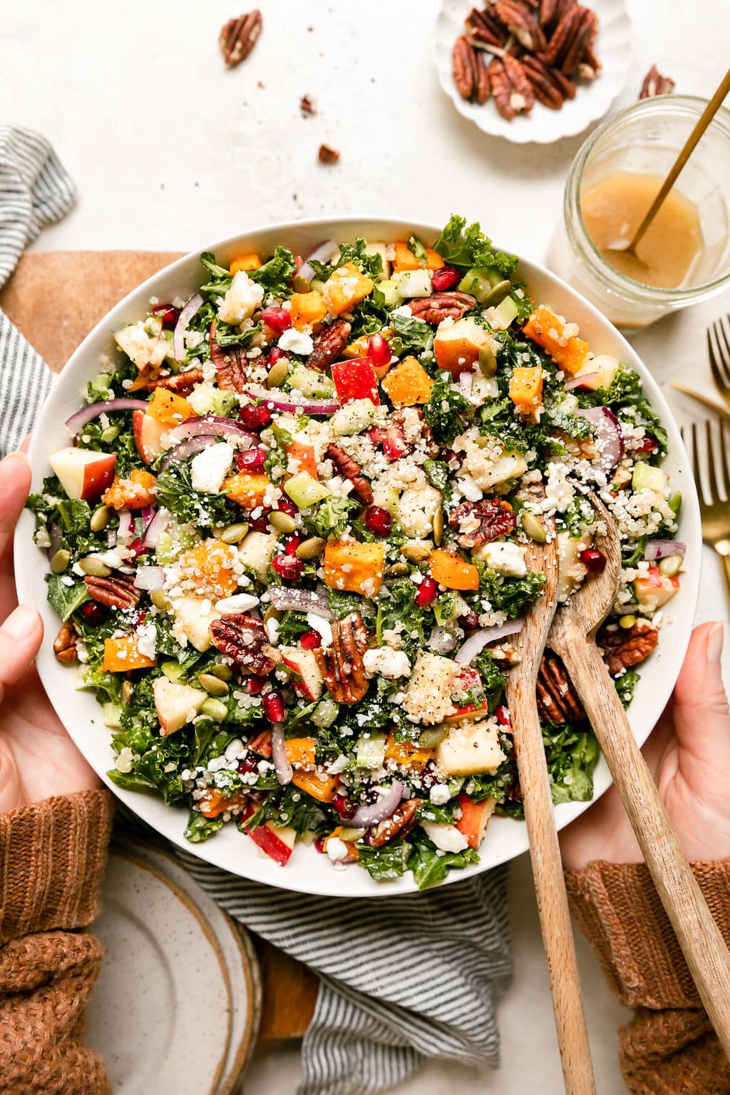 Two hands holding large white serving bowl filled with Harvest Salad with quinoa and butternut quash, wooden serving spoons in salad.