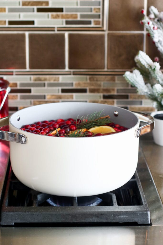 A white pot filled with Christmas Potpourri of fresh cranberries, orange slices, evergreen sprigs and water on a stovetop.