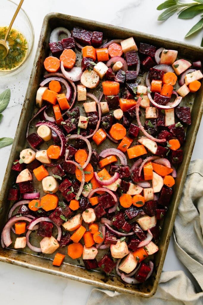 Chopped carrots, beets, sweet potatoes, and parsnips coated in dressing on a baking sheet ready to be roasted.